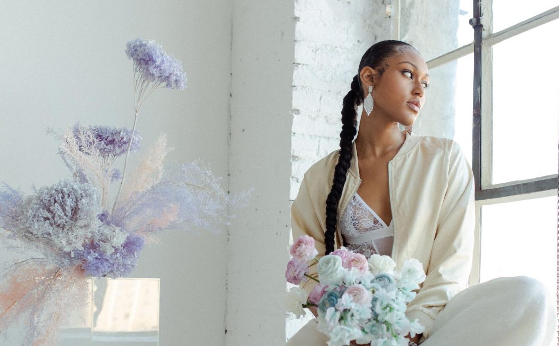 Black bride wearing a cream track suit looks outside of the window while holding a blue and purple bouquet.